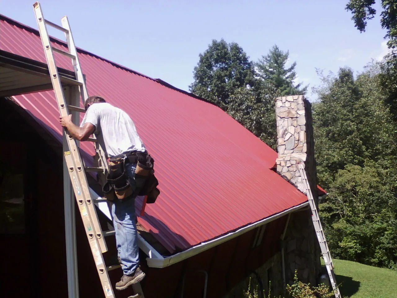 Roofer on a ladder beside a red metal roof and stone chimney — Soffit & Fascia Repair by Roofing Brothers in Sula